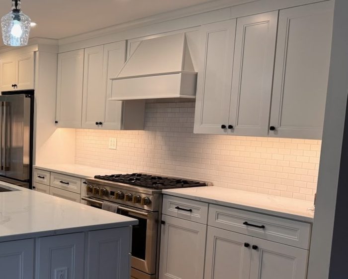 White range hood and shaker cabinets over a stainless gas range, with under-cabinet lighting on a white subway-tile backsplash
